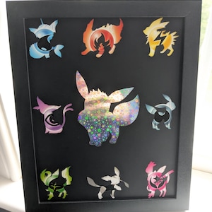 May include: A framed artwork featuring nine colorful, stylized character silhouettes on a black background. The central figure is a sparkly, multi-colored Eevee. Other characters include blue, orange, yellow, purple, silver, green, white, and pink designs. The frame is black.