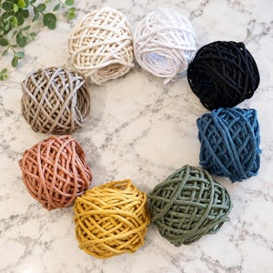 May include: A collection of eight round balls of thick cotton cord arranged in a circle. The colors include cream, white, black, dark blue, olive green, mustard yellow, terracotta, and light brown, all featuring a braided texture.