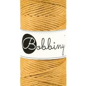 May include: A spool of mustard-colored macrame cord. The cord is tightly wound and wrapped with a white label featuring the word "Bobbiny" in a black oval. The label is dotted along the top and bottom edges.