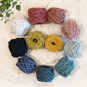May include: A collection of eleven round balls of braided cord arranged in a circular pattern. The colors include white, black, various shades of pink, yellow, and multiple tones of blue, all featuring a textured, woven surface structure.