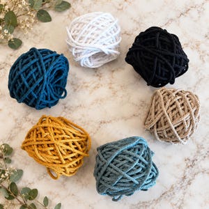 May include: Six balls of thick cord arranged in a circle, featuring colors of white, black, beige, light blue, mustard yellow, and dark teal. Each ball is composed of a continuous length of braided cord wound into a round shape.