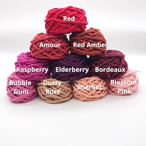 May include: A collection of 10 cotton rope yarn skeins in shades of red, pink, and brown. The skeins are arranged in a pyramid shape. The colors are labeled with names like "Red", "Amour", "Raspberry", "Bubble Gum", "Dusty Rose", "Sherbet", "Blossom Pink", "Bordeaux", "Elderberry", and "Red Amber".