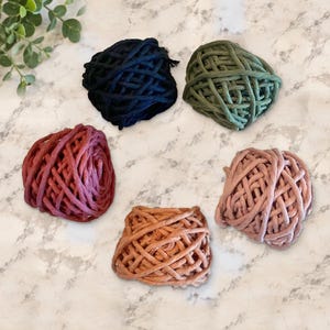 May include: Five bundles of thick, braided cord arranged in a circular pattern. The colors include black, olive green, deep red, light terracotta, and dusty rose. Each bundle consists of multiple strands woven into a loose, textured ball shape.