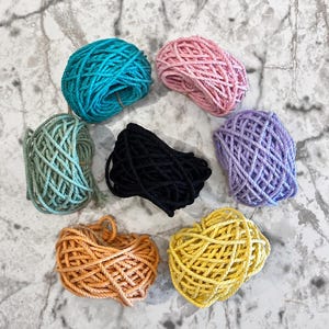 May include: Seven individual bundles of twisted cotton cord arranged in a circular pattern. The colors include teal, light pink, sage green, black, lavender, orange, and yellow. Each bundle consists of multiple strands of rope coiled together.