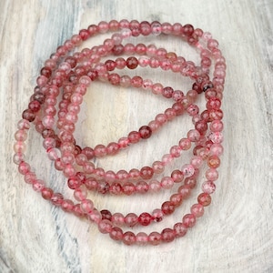 May include: A strand of pink and clear gemstone beads, likely rose quartz, arranged in a circle on a light wood surface.