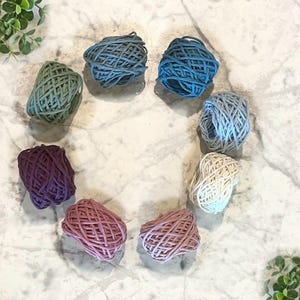 May include: Eight small, round bundles of twine arranged in a circle. The twine colors include sage green, dark blue, medium blue, light gray, cream, dusty rose, mauve, and deep purple. Each bundle features a textured, crisscross pattern.