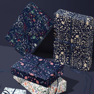 K&ouml;nnte beinhalten: Mehrere Geschenkboxen, verpackt in gemustertem Papier mit floralen Designs in Marineblau, Creme und Rot. Jede Box ist mit einem marineblauen Band gebunden. Die Boxen sind gestapelt und auf einer dunkelblauen Oberfl&auml;che angeordnet.