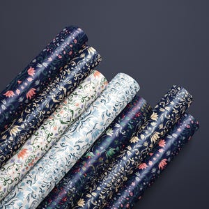 K&ouml;nnte beinhalten: Mehrere Rollen Geschenkpapier mit floralen Mustern in verschiedenen Farben, darunter Marineblau, Wei&szlig; und Rosa. Das Papier zeigt ein sich wiederholendes botanisches Design mit Blumen und Bl&auml;ttern. Die Rollen sind diagonal vor dunklem Hintergrund angeordnet.