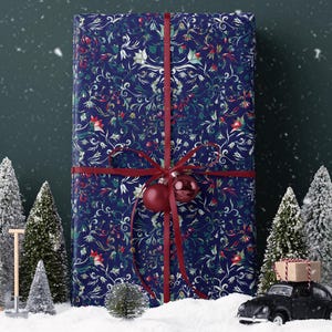 K&ouml;nnte beinhalten: Ein gro&szlig;es, rechteckiges Geschenk, verpackt in marineblauem Papier mit floralem Muster und mit einem roten Band gebunden. Eine rote Kugel h&auml;ngt am Band. Miniatur-schneebedeckte B&auml;ume und ein schwarzes Spielzeugauto mit einem Paket stehen im Vordergrund.