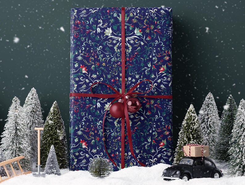 K&ouml;nnte beinhalten: Ein gro&szlig;es, rechteckiges Geschenk, verpackt in marineblauem Papier mit floralem Muster und mit einem roten Band gebunden. Eine rote Kugel h&auml;ngt am Band. Miniatur-schneebedeckte B&auml;ume und ein schwarzes Spielzeugauto mit einem Paket stehen im Vordergrund.