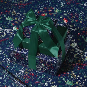 K&ouml;nnte beinhalten: Ein Geschenkbox-Set mit einem dunkelblauen floralen Muster, gekr&ouml;nt von einer gro&szlig;en smaragdgr&uuml;nen Satinschleife. Die Boxen liegen auf passendem floralem Geschenkpapier, was eine zusammenh&auml;ngende und elegante Pr&auml;sentation ergibt.