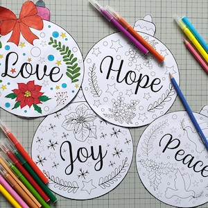 May include: Four Christmas ornament-shaped coloring pages with the words "Love", "Hope", "Joy", and "Peace". Each ornament has a unique design with stars, leaves, and flowers. Colored markers and pencils are scattered around the ornaments.