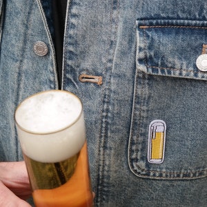 Könnte beinhalten: Eine blaue Jeansjacke mit einem weißen Kunstfellkragen. Die Jacke hat eine Tasche auf der linken Seite mit einer kleinen Metallstifte mit einem Bierglas. Die Person hält ein Glas Bier.