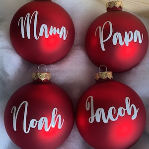 May include: Four red Christmas ornaments with gold caps, each inscribed with a white name: "Mama", "Papa", "Noah", and "Jacob". The ornaments are arranged in a square, resting on a bed of white fluff, suggesting a holiday theme.