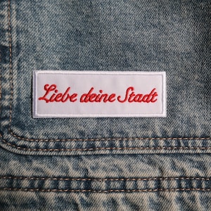 Könnte beinhalten: Nahaufnahme eines Jeansstoffs mit einer Tasche. Auf der Tasche ist ein weißes rechteckiges Patch mit dem rot gestickten Schriftzug "Liebe deine Stadt" aufgenäht. Der Denim hat eine verwaschene blaue Waschung und sichtbare Nähte.
