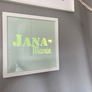 May include: A framed artwork with the name "JANA-Marie" in a neon green font against a light green background. The frame is white and mounted on a gray wall. A decorative branch is visible on the right side of the image.