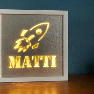 Könnte beinhalten: Ein weißer Rahmen mit silbernem Glitzerhintergrund. Der Rahmen hat ein ausgeschnittenes Design eines Raumschiffs und den Namen "MATTI" in goldenen Buchstaben.