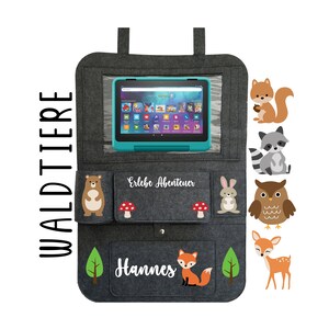May include: Gray felt car seat organizer with pockets and a clear window for a tablet. The organizer has a white inscription that says "Erlebe Abenteuer" and "Hannes". The organizer has several colorful animal illustrations, including a squirrel, raccoon, owl, deer, bear, and fox.