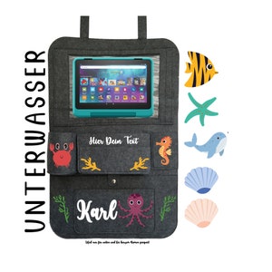 May include: Gray felt tablet holder with a clear plastic window for a tablet. The holder has pockets on the sides and a pocket on the bottom. The pockets are decorated with sea creatures, including a crab, a seahorse, an octopus, a starfish, a whale, and a fish. The text "Hier Dein Text" is printed on the holder. The text "Karl" is printed on the bottom pocket. The text "Ideal f&uuml;r Auto und f&uuml;r zu Hause. Montage m&ouml;glich" is printed on the bottom of the holder.