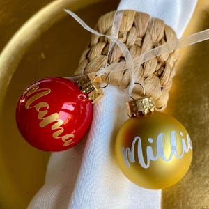 May include: Two Christmas ornaments, one red and one gold, with names in cursive script. The red ornament says "Mama" and the gold ornament says "Millie". Both are attached to a woven napkin ring with white ribbon.