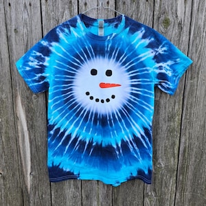 Może przedstawiać: Koszulka w kolorze niebieskim i białym z tie-dye z wzorem białej twarzy bałwana. Bałwan ma czarne oczy, marchewkowy nos i usta.