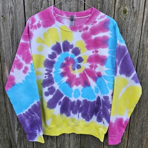 Może przedstawiać: Bluza z okrągłym dekoltem z żywym wzorem tie-dye. Wzór przedstawia spiralę kolorów, w tym różowy, żółty, niebieski i fioletowy. Bluza wisi na tle zwietrzałego drewnianego tła.