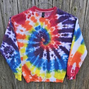 Może przedstawiać: Bluza z efektem tie-dye w spiralny wzór, z żywą mieszanką kolorów, w tym czerwonym, pomarańczowym, żółtym, niebieskim i fioletowym. Bluza ma okrągły dekolt i długie rękawy z żółtymi mankietami. Odzież wisi na srebrnym wieszaku.
