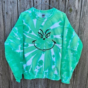 May include: Green and white tie-dye sweatshirt with a black Grinch face design. The sweatshirt is suspended on a metal hanger against a wooden backdrop. The Grinch face features eyes and a smile.