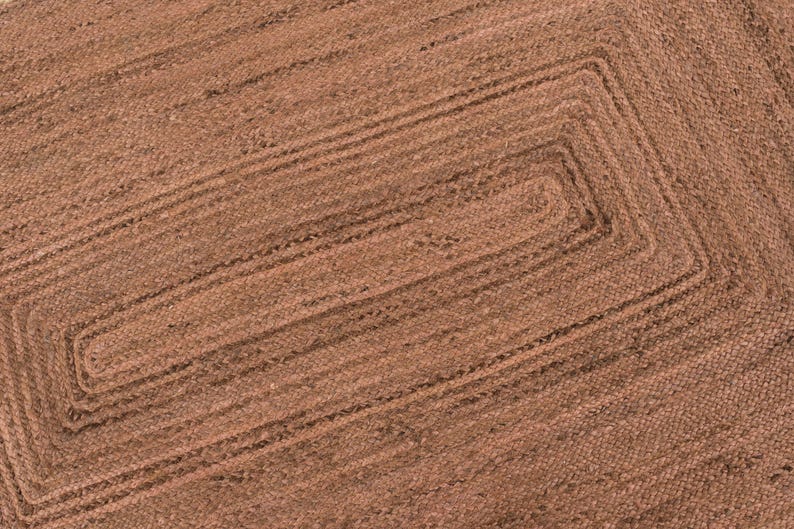 Op de afbeelding: Close-up van een rechthoekig bruin jute vloerkleed met een geweven ontwerp. Het vloerkleed heeft een centraal rechthoekig patroon met concentrische randen. De natuurlijke vezels cre&euml;ren een textuuroppervlak.