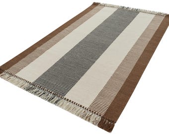 May include: Rectangular rug with a striped pattern in brown, cream, and black. The rug has a fringed edge and is made of woven material. The design features alternating wide and narrow stripes, creating a simple yet stylish look.