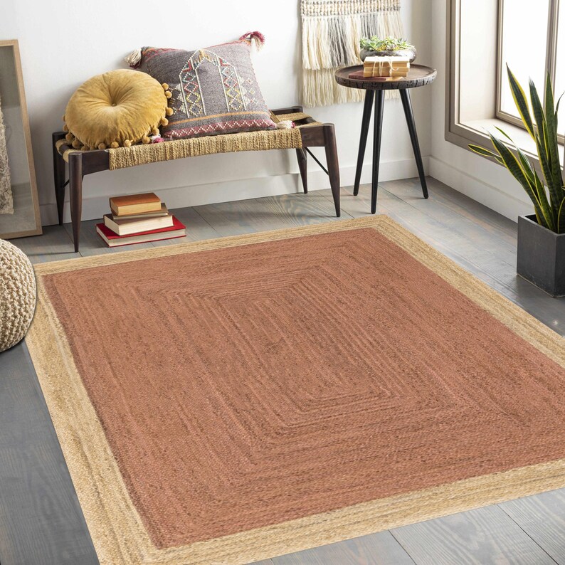 Op de afbeelding: Een rechthoekig jute vloerkleed met een vierkant ontwerp in het midden. Het vloerkleed heeft een lichtbruine rand en een roodbruin midden. Het vloerkleed ligt op een houten vloer, met een bank en kussens op de achtergrond.
