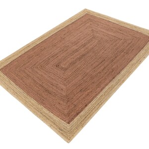 Op de afbeelding: Rechthoekig jute vloerkleed met een natuurlijke beige rand en een middenstuk in een roodbruine tint. Het vloerkleed heeft een geometrisch ontwerp met een reeks concentrische rechthoeken. Het vloerkleed is gemaakt van geweven jutevezels.