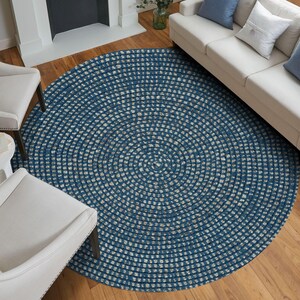 Pode incluir: Um tapete redondo tran&ccedil;ado azul e bege. O tapete &eacute; feito de material tecido e tem um padr&atilde;o circular. O tapete &eacute; colocado em um piso de madeira, com um sof&aacute; branco e cadeiras ao fundo.