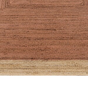 Op de afbeelding: Close-up van een geweven jute vloerkleed met een vierkant patroon. Het vloerkleed heeft een warme, roodbruine kleur in het midden, omlijst door een lichtere beige rand. De textuur is zichtbaar en toont de natuurlijke vezels en de constructie van het vloerkleed.