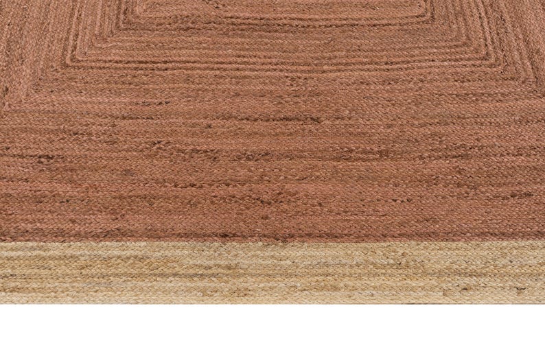 Op de afbeelding: Close-up van een geweven jute vloerkleed met een vierkant patroon. Het vloerkleed heeft een warme, roodbruine kleur in het midden, omlijst door een lichtere beige rand. De textuur is zichtbaar en toont de natuurlijke vezels en de constructie van het vloerkleed.