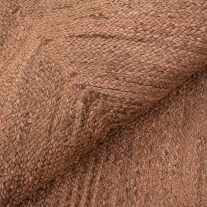 Op de afbeelding: Close-up van een geweven tapijt met een natuurlijke, aardse tint. De textuur van het tapijt is zichtbaar, met een patroon van verweven vezels. De kleur is een warm, roodbruin, wat wijst op een rustieke of bohemien stijl. Het tapijt is gedeeltelijk opgerold.