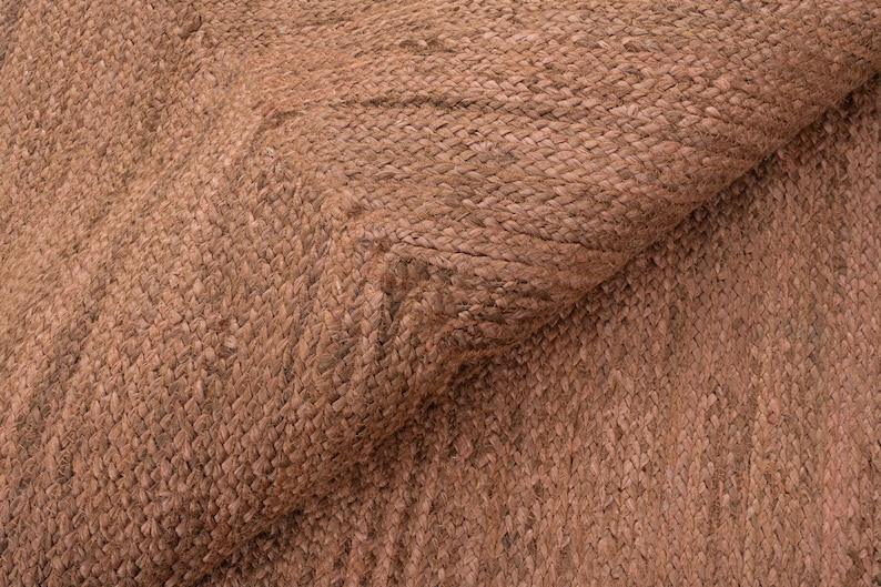 Op de afbeelding: Close-up van een geweven tapijt met een natuurlijke, aardse tint. De textuur van het tapijt is zichtbaar, met een patroon van verweven vezels. De kleur is een warm, roodbruin, wat wijst op een rustieke of bohemien stijl. Het tapijt is gedeeltelijk opgerold.