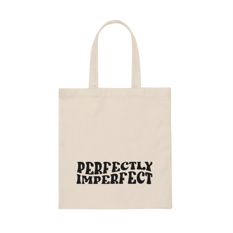 Peut inclure: Un sac fourre-tout en toile beige avec du texte noir qui dit "PERFECTLY IMPERFECT".