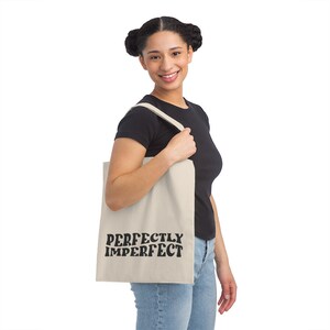 Peut inclure: Une personne tient un sac fourre-tout beige avec du texte noir qui dit "PERFECTLY IMPERFECT".