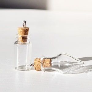 May include: Two small, clear glass bottles with cork stoppers. One bottle stands upright, while the other lies on its side. Each bottle has a small metal loop attached to the cork. The bottles are on a white surface.