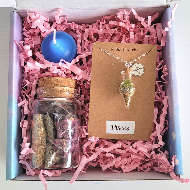 May include: A gift box containing pink paper shred, a blue candle, a glass jar with a cork, and a necklace with a glass vial pendant. The necklace has a Pisces charm and the text "RJSpellworks" is visible. The label reads "Pisces".