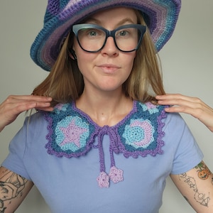 K&ouml;nnte beinhalten: Eine Frau tr&auml;gt einen lilafarbenen H&auml;kelhut mit breiter Krempe und einen lilafarbenen H&auml;kelkragen mit blauen und rosa Akzenten. Der Kragen hat ein Sternmuster und eine Schleife.