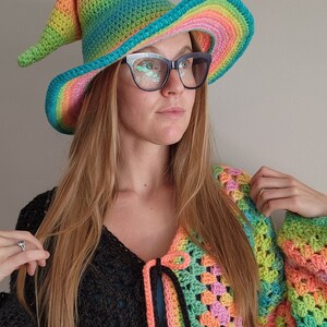 K&ouml;nnte beinhalten: Eine Frau tr&auml;gt einen Regenbogen-H&auml;kelhut mit breiter Krempe und eine passende Regenbogen-H&auml;keljacke. Der Hut hat eine spitze Krone und eine lange, flie&szlig;ende Krempe.