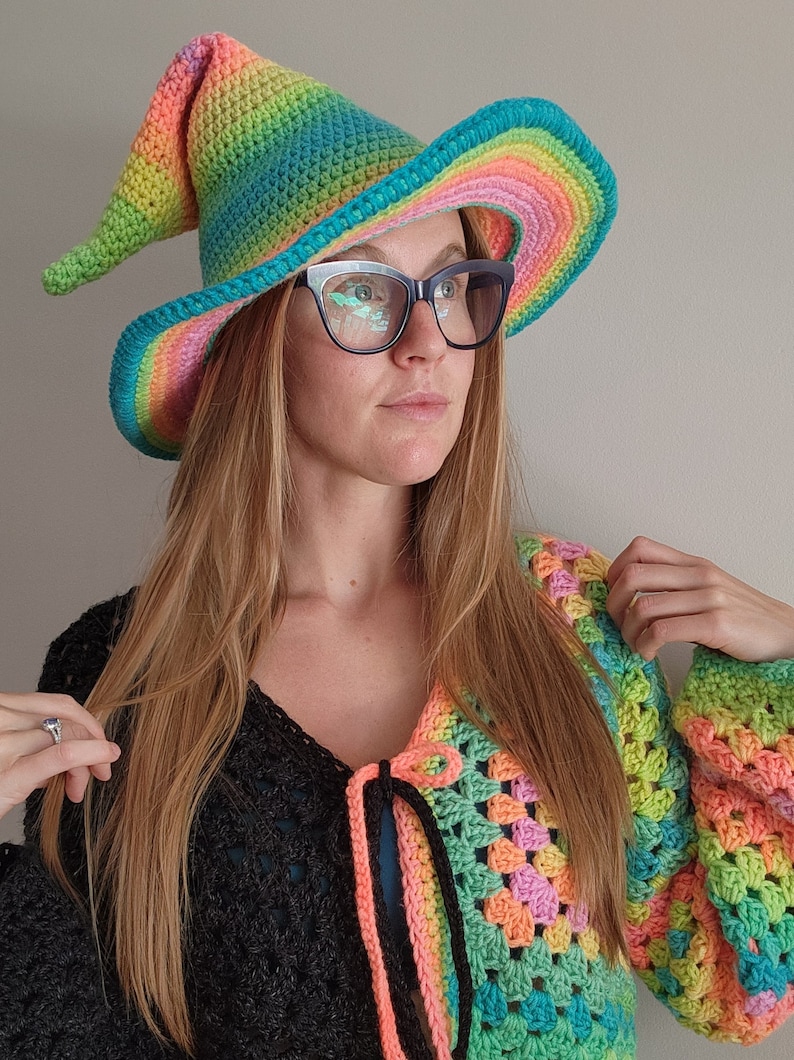 K&ouml;nnte beinhalten: Eine Frau tr&auml;gt einen Regenbogen-H&auml;kelhut mit breiter Krempe und eine passende Regenbogen-H&auml;keljacke. Der Hut hat eine spitze Krone und eine lange, flie&szlig;ende Krempe.