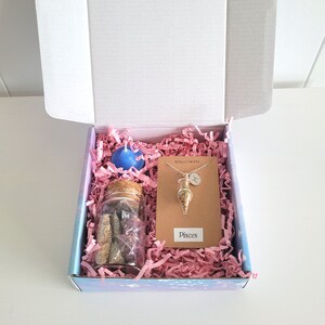 May include: A gift box containing a blue candle, a glass jar with a cork lid filled with items, and a Pisces necklace. The box is lined with pink paper and has a label that says "Pisces".