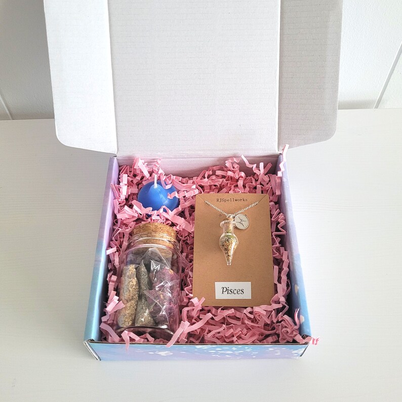 May include: A gift box containing a blue candle, a glass jar with a cork lid filled with items, and a Pisces necklace. The box is lined with pink paper and has a label that says "Pisces".