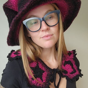 K&ouml;nnte beinhalten: Eine Frau tr&auml;gt ein schwarzes Oberteil mit einem rosa und schwarzen H&auml;kelkragen und einen breitkrempigen rosa und schwarzen H&auml;kelhut. Sie tr&auml;gt auch eine schwarze Brille.