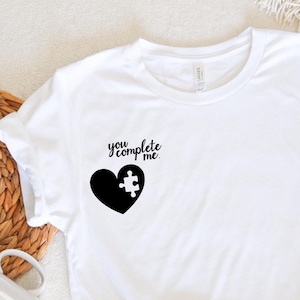May include: White t-shirt with a black heart and puzzle piece design. The text "you complete me" is written above the heart.