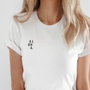 Può includere: Una maglietta bianca con la scritta "ALOHA." stampata sul davanti.