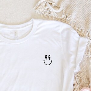 Op de afbeelding: Wit T-shirt met een zwarte smiley-print. De smiley heeft twee zwarte stippen voor ogen en een gebogen lijn voor een mond.